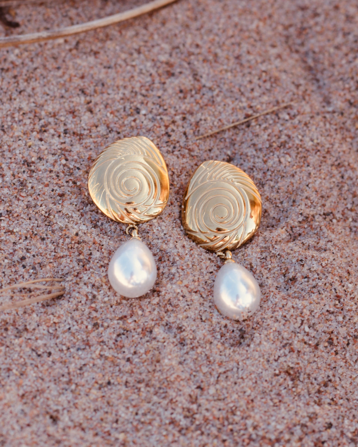 A pair of gold drop earrings with a spiral patterned top and a dangling white pearl, resting on a sandy surface.
